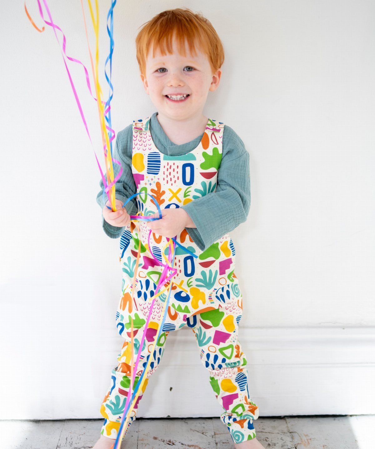 Red-haired toddler girl in colorful patterned overalls holding bright curly streamers against a white wall.