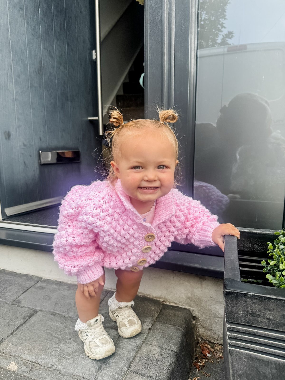 Smiling toddler girl wearing a chunky pink knit cardigan and white sneakers standing outdoors.