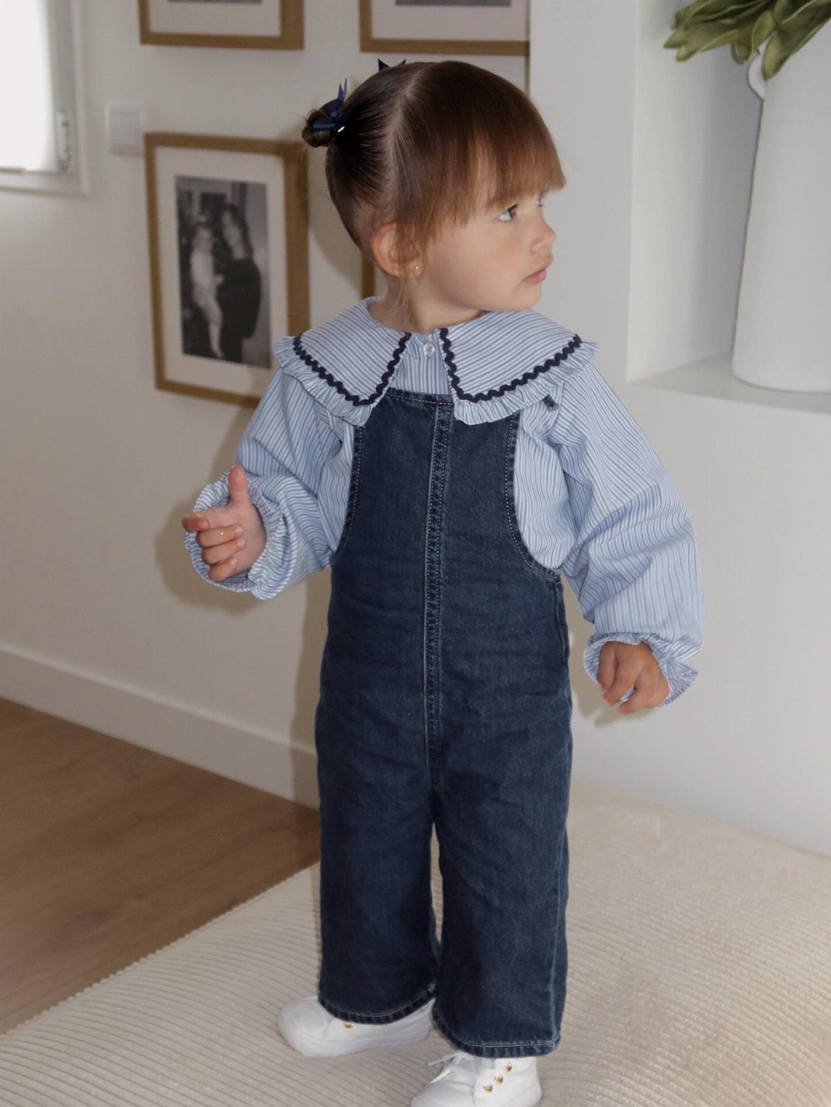 Toddler girl wearing denim overalls over a blue striped shirt and white sneakers.