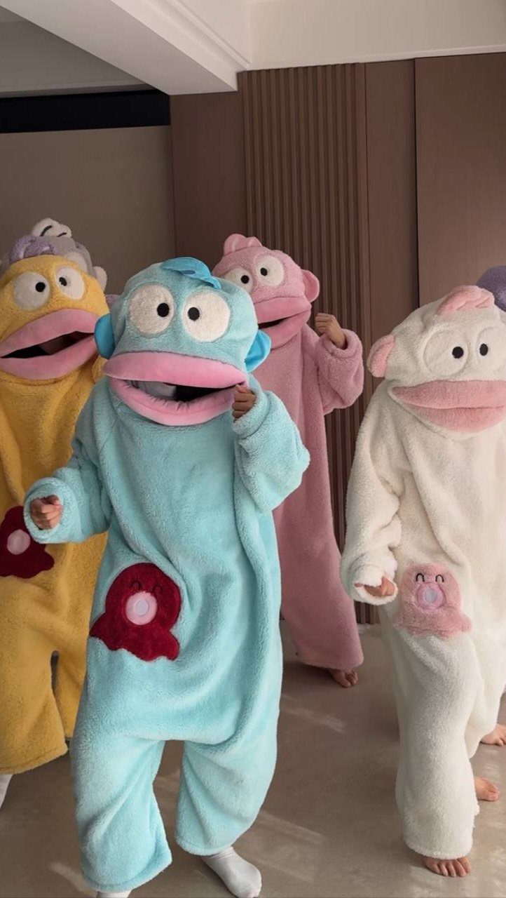 Four toddlers wearing colorful plush character fleece onesies standing indoors.