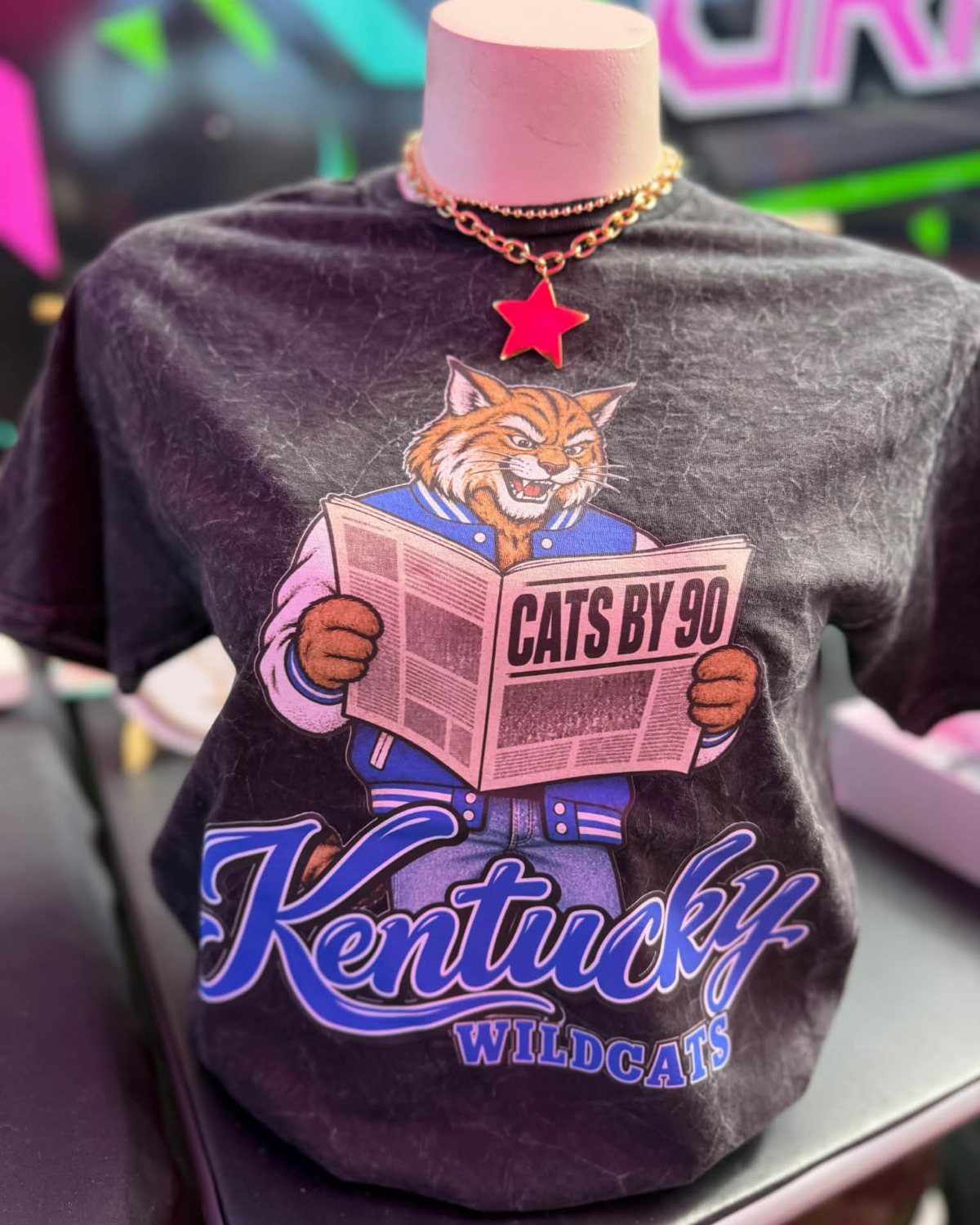 A black Kentucky Wildcats graphic t-shirt displayed on a pink mannequin torso with a star necklace.