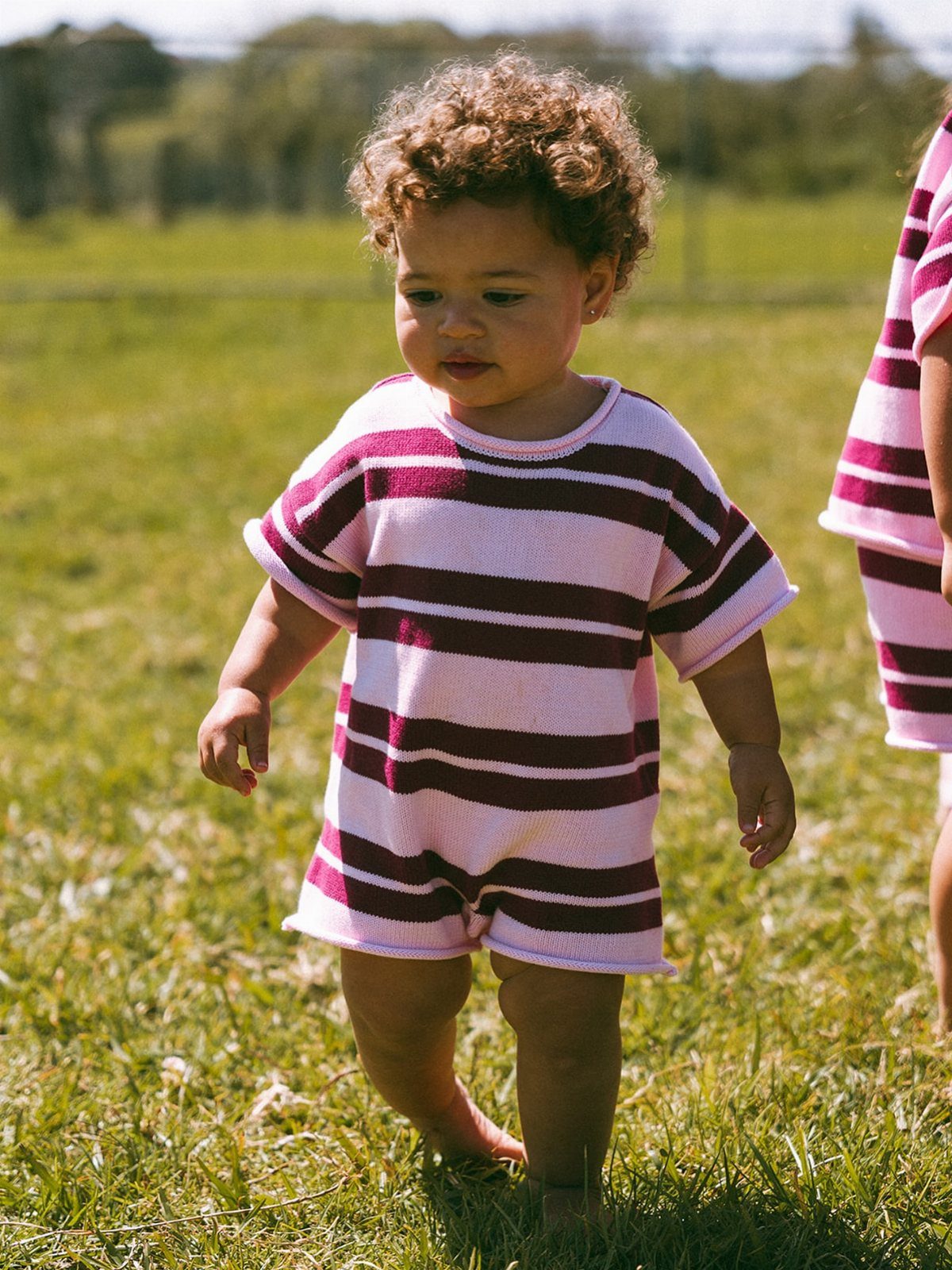 Toddler girl with curly hair wearing a pink and maroon striped romper walking on grass.