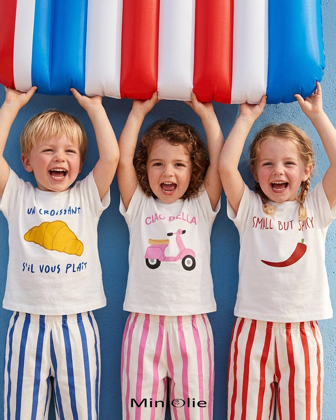 Three laughing toddlers holding a large striped inflatable against a bright blue wall.